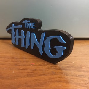 以下が含まれることがあります： スタイリッシュなフォントで「THE THING」という文字が書かれた、黒と青の3Dプリントされた看板です。