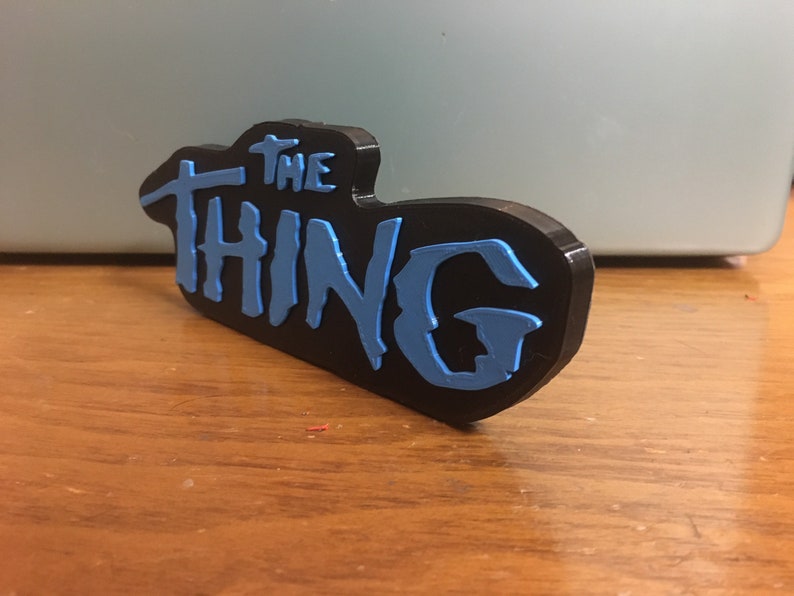 以下が含まれることがあります： スタイリッシュなフォントで「THE THING」という文字が書かれた、黒と青の3Dプリントされた看板です。