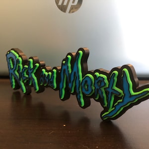 Może przedstawiać: Tr&oacute;jwymiarowa tabliczka z napisem "Rick and Morty" w stylizowanym kroju pisma. Tabliczka jest czarna z niebieskimi i zielonymi akcentami.