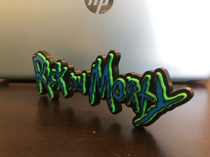 Może przedstawiać: Tr&oacute;jwymiarowa tabliczka z napisem "Rick and Morty" w stylizowanym kroju pisma. Tabliczka jest czarna z niebieskimi i zielonymi akcentami.