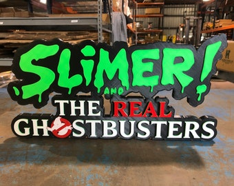Pode incluir: Uma placa 3D preta e verde com as palavras "Slimer! And The Real Ghostbusters" em letras brancas, vermelhas e verdes. A placa é feita de espuma e tem uma superfície texturizada.