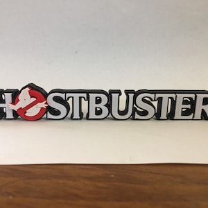 May include: A black and white plastic sign with the word "GHOSTBUSTERS" in white letters with a red and white ghost logo in the middle.