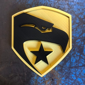 May include: A gold-colored shield with a black eagle and a black five-pointed star. The eagle has a yellow eye.