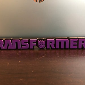 May include: Purple "TRANSFORMERS" logo with the Autobot symbol in the middle. The logo is made of plastic and is sitting on a wooden surface.