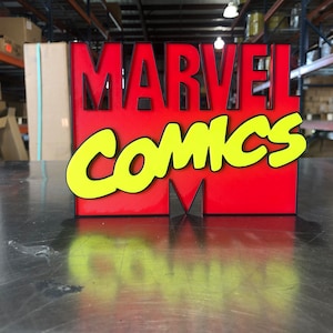 May include: A red and yellow 3D sign with the words "MARVEL COMICS" in a bold, stylized font. The sign is on a metal surface.