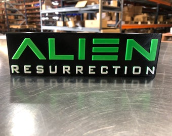 Pode incluir: Uma placa impressa em 3D preta e verde com o texto "ALIEN RESURRECTION" em letras brancas.
