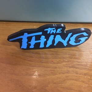 以下が含まれることがあります： 黒い3Dプリントされたサインに青い文字で「THE THING」と書かれている
