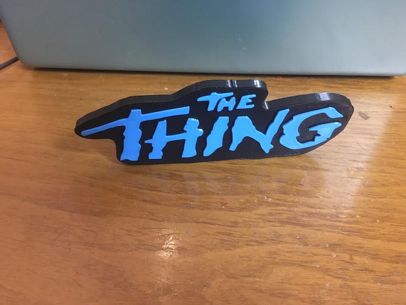以下が含まれることがあります： 黒い3Dプリントされたサインに青い文字で「THE THING」と書かれている