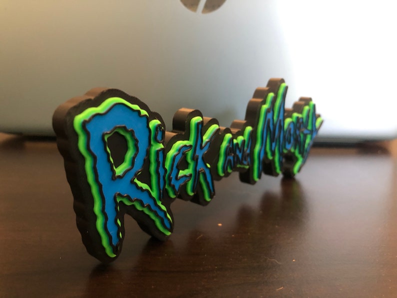Może przedstawiać: Tr&oacute;jwymiarowy znak z napisem "Rick and Morty" w kolorze niebieskim, zielonym i czarnym.