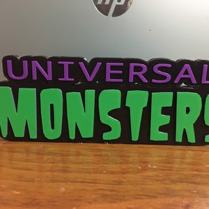May include: A 3D printed sign with the words "Universal Monsters" in green and purple letters on a black background.