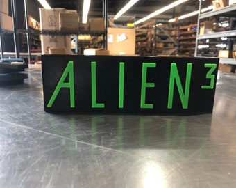 Pode incluir: Uma placa preta com as palavras "ALIEN 3" em letras verdes.