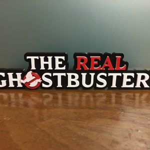 May include: A black plastic sign with the words "THE REAL GHOSTBUSTERS" in white letters. The word "REAL" is in red letters. A white ghost with a red no symbol is in front of the word "GHOSTBUSTERS".