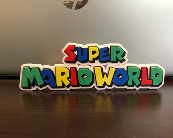 Pode incluir: Um emblema retangular e colorido do Super Mario World com uma borda branca. O texto "SUPER" é em vermelho, azul e amarelo, enquanto "MARIO WORLD" é em verde, amarelo, azul e vermelho. O emblema está sobre uma superfície marrom escura.
