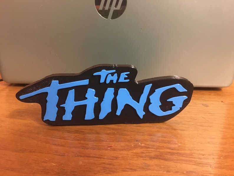 以下が含まれることがあります： スタイリッシュなフォントで「THE THING」と書かれた黒と青の3Dプリントサイン