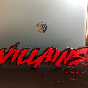 May include: A 3D printed sign that says "VILLAINS" in red and black letters. The sign is on a black surface with red splatters.