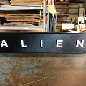 May include: A black rectangular sign with the word "ALIEN" in white raised letters.