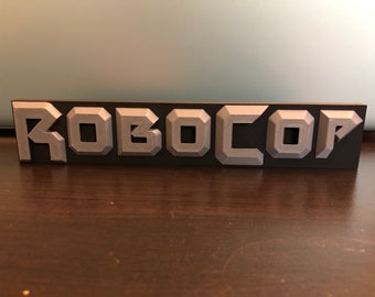 Puede incluir: Una placa negra con la palabra "ROBOCOP" en letras plateadas en relieve.