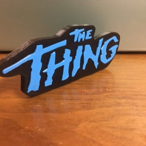 以下が含まれることがあります： 黒い3Dプリントされたサインに青い文字で「THE THING」と書かれている