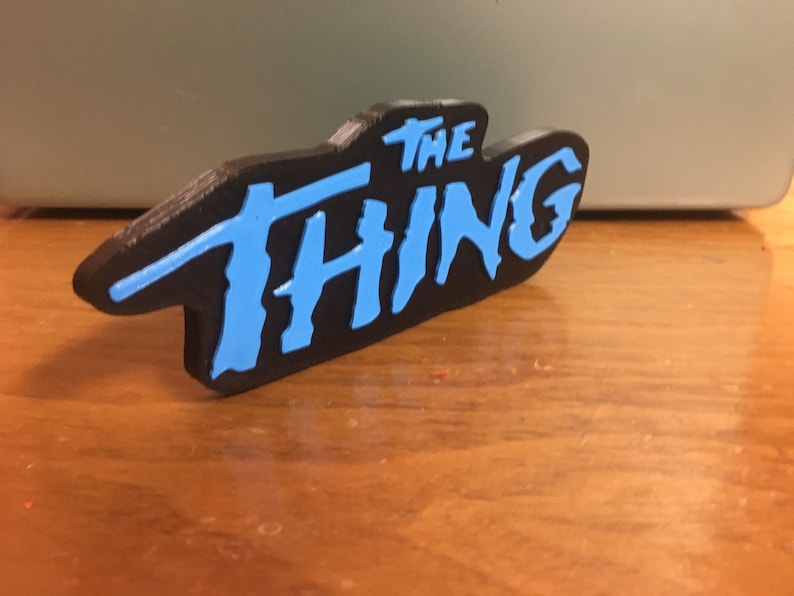 以下が含まれることがあります： 黒い3Dプリントされたサインに青い文字で「THE THING」と書かれている