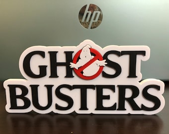Pode incluir: Um sinal branco impresso em 3D com as palavras "Ghostbusters" em letras pretas. Um círculo vermelho com uma linha diagonal que o atravessa e um fantasma branco dentro está centrado entre as palavras "Ghost" e "busters".