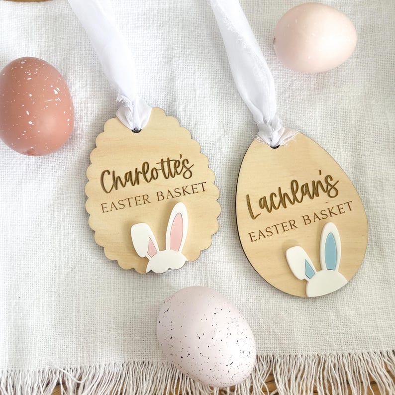 May include: Two wooden Easter basket tags with bunny ears. One tag is oval shaped and says "Lachlan's Easter Basket" with blue bunny ears. The other tag is round and says "Charlotte's Easter Basket" with pink bunny ears.
