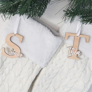 May include: Two white knitted Christmas stockings with wooden ornaments. The ornaments are shaped like the letters S and T, with the names "Sonny" and "Thea" written on them.