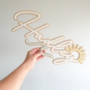 May include: A wooden sign spelling "Hadley" in a layered design. The sign has a light brown base with white and light blue accents, and a yellow sun graphic. The sign is held by a person's hand.