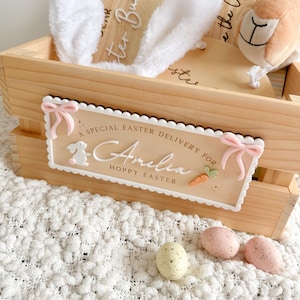 May include: A wooden Easter crate with a sign reading "A Special Easter Delivery For Amelia Hoppy Easter." The crate holds white bunny ears, a plush lamb, and a wooden sign. Three decorative eggs are in front.