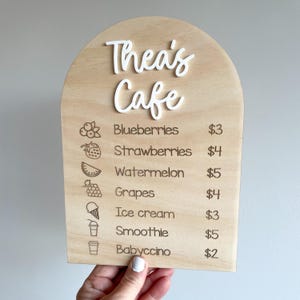 May include: Wooden cafe menu board with the text "Thea's Cafe" in white script. The menu lists blueberries, strawberries, watermelon, grapes, ice cream, smoothie, and babyccino with prices.
