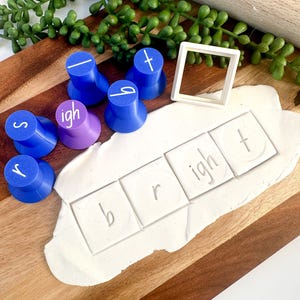 May include: A set of blue and purple plastic letter stamps and a white square frame cutter. The stamps are used to press the letters b, r, i, g, h, and t into a piece of white modelling clay, forming the word bright on the surface of the clay.