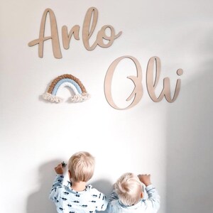 May include: Two children stand in front of a white wall with wooden name signs that read "Arlo" and "Oli". A small rainbow-coloured macrame wall hanging is above the names.