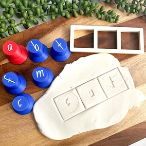May include: A set of plastic letter stamps in red and blue with white lowercase letters, a white rectangular three-section frame, and a piece of white modelling dough featuring the impressed letters c, a, and f within square outlines.