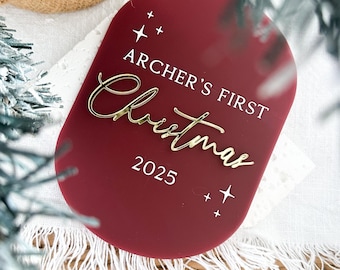 May include: A burgundy acrylic Christmas ornament with the text "ARCHER'S FIRST Christmas 2025" in white and gold. The ornament has a rounded rectangular shape and star accents. The ornament is placed on a white surface with a woven basket in the background.
