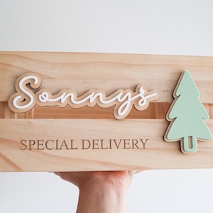 May include: A wooden crate with the words "Sonny's Special Delivery" engraved on the front. The crate is decorated with a white and brown cursive name and a green paper cutout of a Christmas tree.