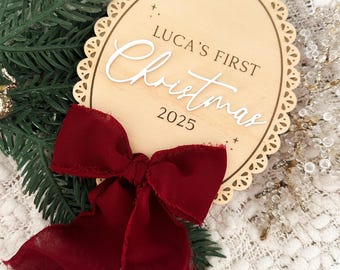 May include: A wooden Christmas ornament with the text "LUCA'S FIRST Christmas 2025" in white script. The ornament has a scalloped edge and is adorned with a deep red bow. Greenery and decorative accents surround the ornament.
