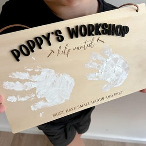 May include: Wooden sign with the words "POPPY'S WORKSHOP" in black 3D letters. The sign also has the words "help wanted" and two white handprints. The bottom of the sign reads "MUST HAVE SMALL HANDS AND FEET".