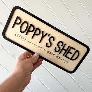 May include: A rectangular wooden sign with the words "POPPY'S SHED" in bold black letters. Below, the text reads "LITTLE HELPERS ALWAYS WANTED". The sign has a black border and a light wood grain background.