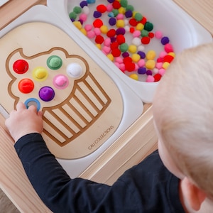 May include: A child is playing with a wooden toy featuring a cupcake design with colourful pom-poms. The toy has a separate compartment filled with colourful pom-poms. The toy is labelled "CAU COLLECTIONS".