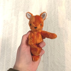 Puede incluir: Un pequeño juguete de peluche de zorro naranja con orejas forradas de blanco y una nariz negra. El zorro se sostiene en una mano sobre un fondo blanco. El juguete tiene una textura suave y peluda y está diseñado para parecerse a un zorro.