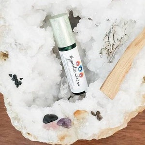 May include: A white geode with a green and white bottle of "Hypnotic Jewels"  essential oil, a piece of sage, a wooden stick, and various crystals.