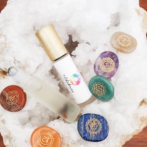 May include: Seven chakra crystal stones in various colors with gold symbols on a white geode. A clear glass bottle with a gold cap and a label that reads "7 Chakras" is in the center of the image.