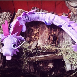 May include: A purple plush dragon toy with a long tail and a large head with pink ears. The dragon is lying on a pile of wood.