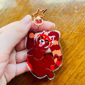 K&ouml;nnte beinhalten: Ein Schl&uuml;sselanh&auml;nger mit einem l&auml;chelnden roten Panda, mit rotem K&ouml;rper und wei&szlig;en Akzenten. Er hat einen sternf&ouml;rmigen Verschluss. Der Schl&uuml;sselanh&auml;nger ist aus klarem Acryl gefertigt.