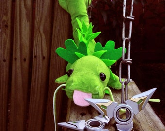 May include: A green plush dragon toy with a pink tongue and two silver fidget spinners rest on a wooden surface. The dragon is hanging from a metal chain. The background is a wooden fence.