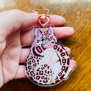 K&ouml;nnte beinhalten: Ein klarer Acryl-Schl&uuml;sselanh&auml;nger mit einem Cartoon-Leoparden-Design. Der Leopard hat einen wei&szlig;en K&ouml;rper, rosa Ohren und ein gepunktetes Muster in Braun- und Lilat&ouml;nen. Der Schl&uuml;sselanh&auml;nger hat einen ros&eacute;goldenen sternf&ouml;rmigen Verschluss.