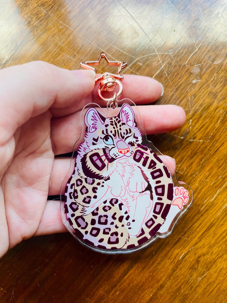 K&ouml;nnte beinhalten: Ein klarer Acryl-Schl&uuml;sselanh&auml;nger mit einem Cartoon-Leoparden-Design. Der Leopard hat einen wei&szlig;en K&ouml;rper, rosa Ohren und ein gepunktetes Muster in Braun- und Lilat&ouml;nen. Der Schl&uuml;sselanh&auml;nger hat einen ros&eacute;goldenen sternf&ouml;rmigen Verschluss.