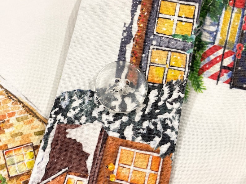 May include: A white fabric with a Christmas-themed print featuring houses with windows and snow. A clear plastic button is visible in the center of the image.
