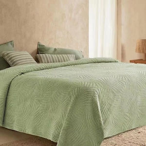May include: A green quilted bedspread with a leaf pattern. The bedspread is on a bed with two pillows and a beige rug.