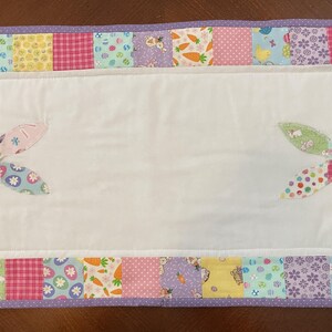 May include: A white Easter-themed table runner with a patchwork border featuring pastel patterns of eggs, carrots, and bunnies. Appliqued bunny ears are on the white center. The border is trimmed with a purple dotted fabric.
