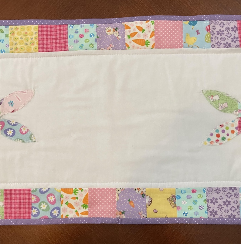 May include: A white Easter-themed table runner with a patchwork border featuring pastel patterns of eggs, carrots, and bunnies. Appliqued bunny ears are on the white center. The border is trimmed with a purple dotted fabric.